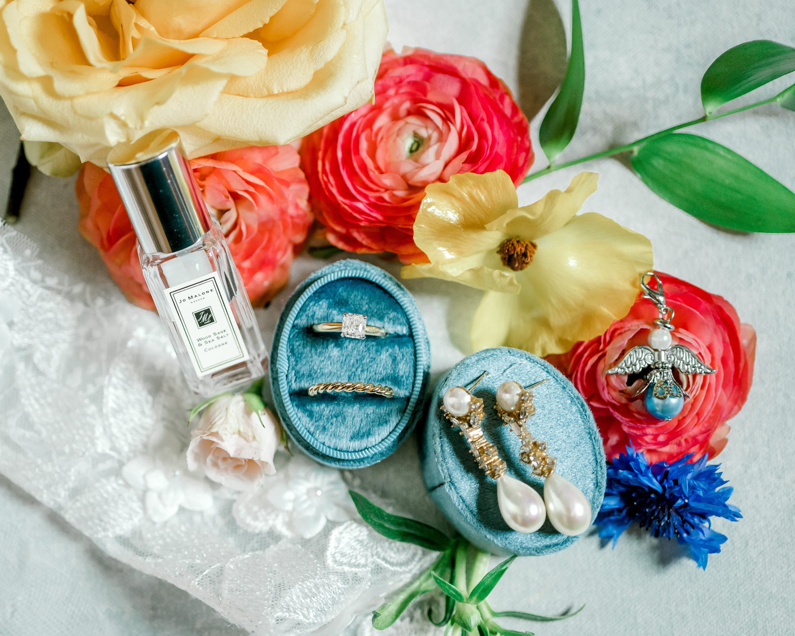 bridal engagement and wedding rings, earrings, and perfume flatlay with bright colorful summer flowers
